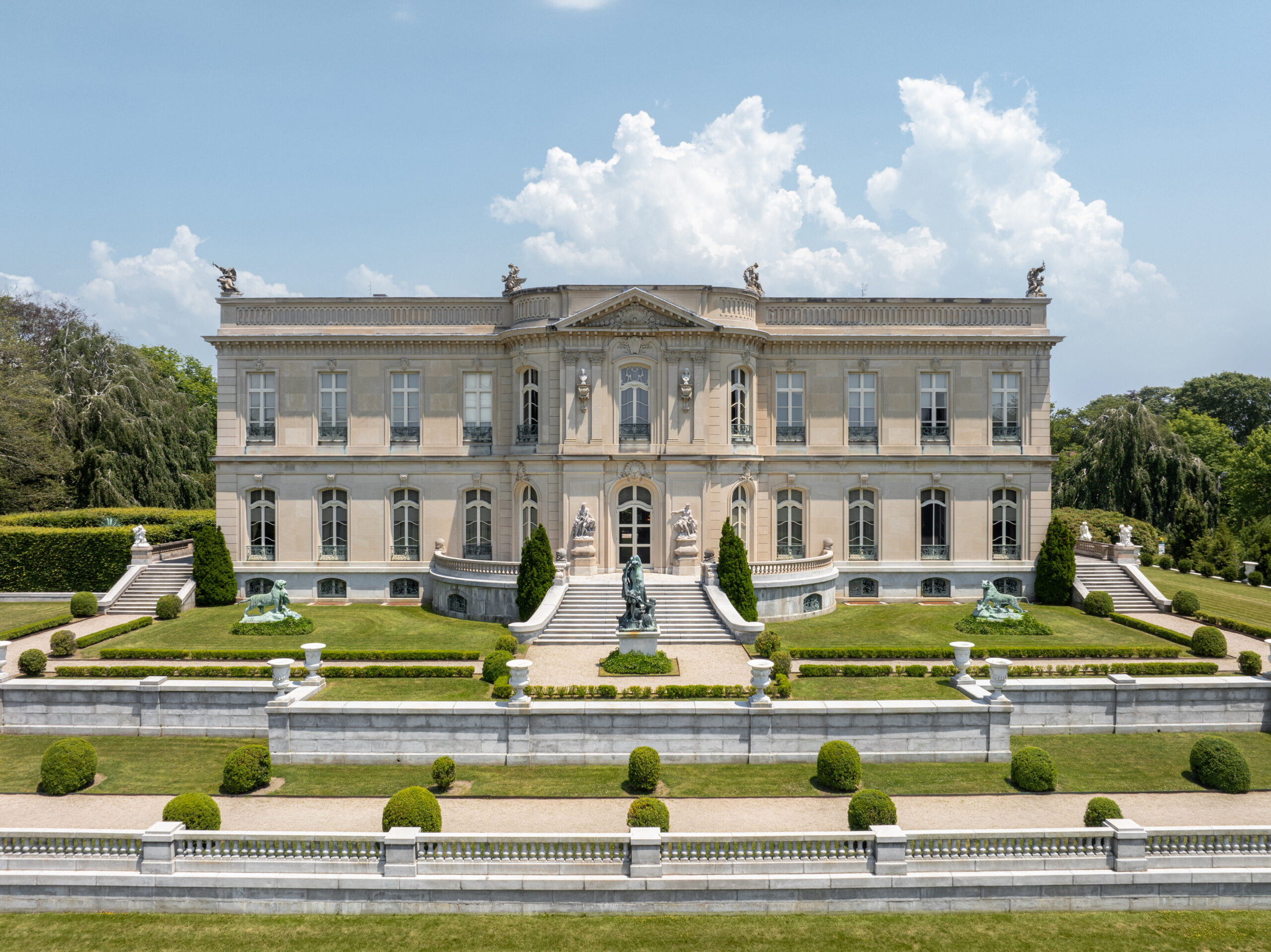 Symmetrical white mansion with statues, trimmed round shrubs, and manicured grass in front