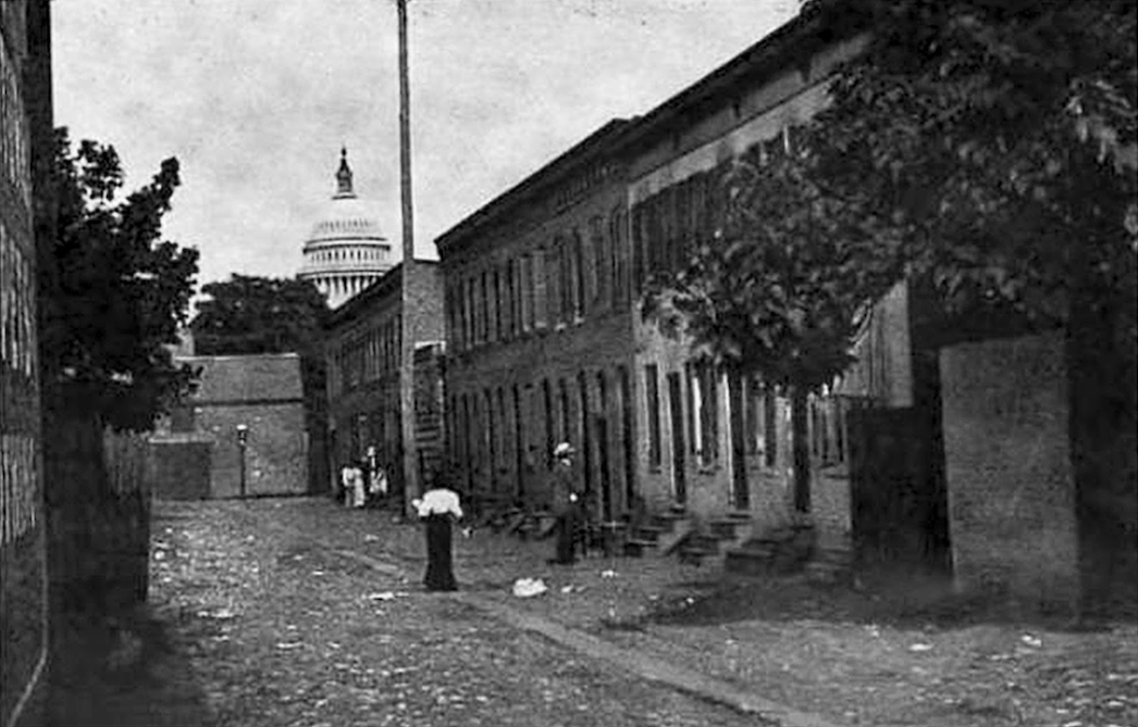 Black and white photograph of people standing in an alley with Capitol Building dome in background