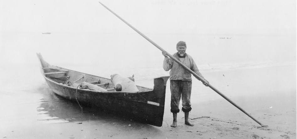 A man holding a pole standing next to a canoe on the beach