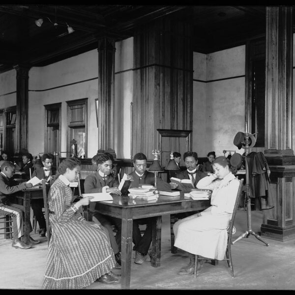 Black and white photograph of interior view of library reading room with male and female students sitting at tables, reading